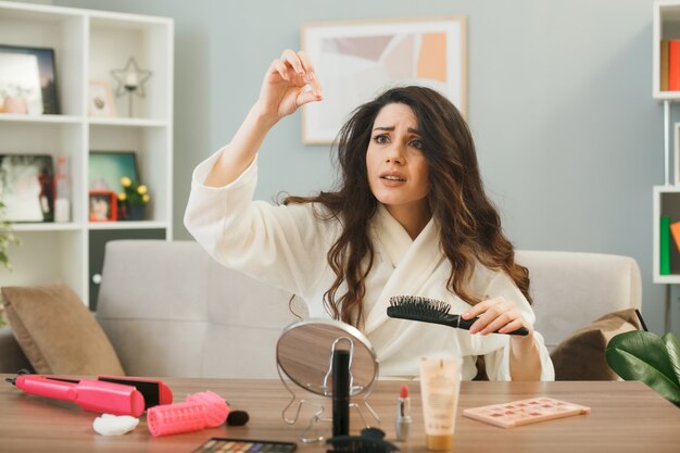 Young woman holding comb sitting at table with makeup tools in living room