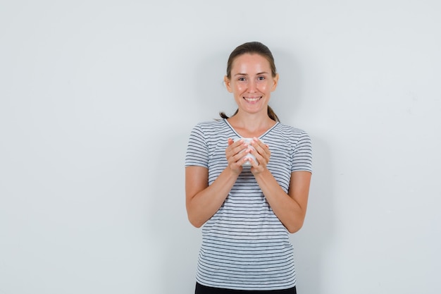 Free photo young woman holding cup of tea in t-shirt, pants and looking cheery. front view.