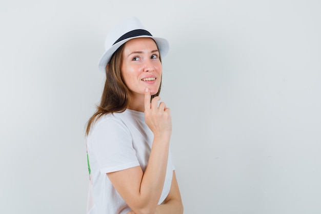 Young woman holding finger on chin in white t-shirt, hat and looking dreamy.