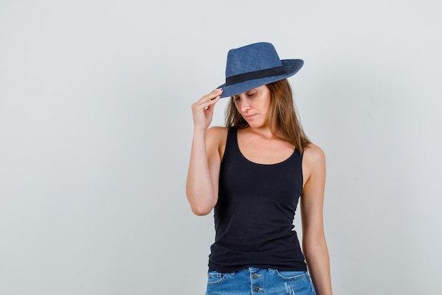 Young woman holding fingers on hat while posing in singlet, shorts and looking charming. front view.