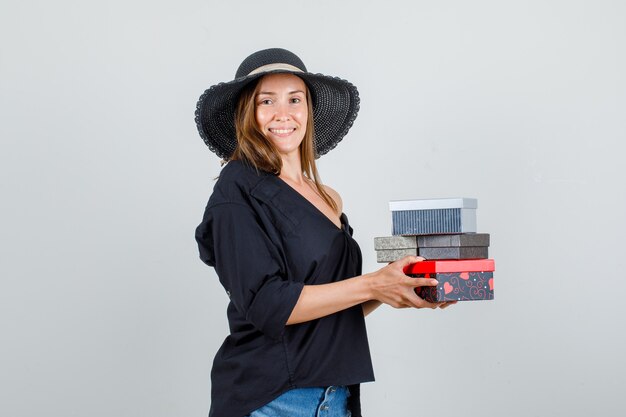 Young woman holding gift boxes in shirt, shorts, hat and looking cheery.