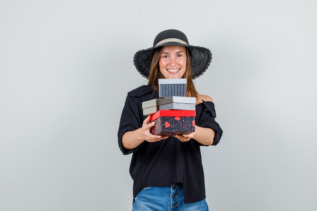 Free photo young woman holding gift boxes in shirt, shorts, hat and looking merry. front view.