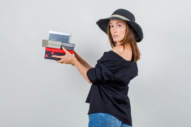 Young woman holding gift boxes while looking back in shirt, shorts, hat .