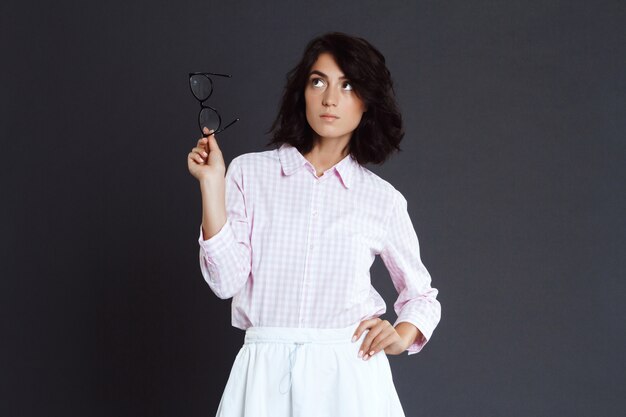 Young woman holding glasses in hand posing over grey wall