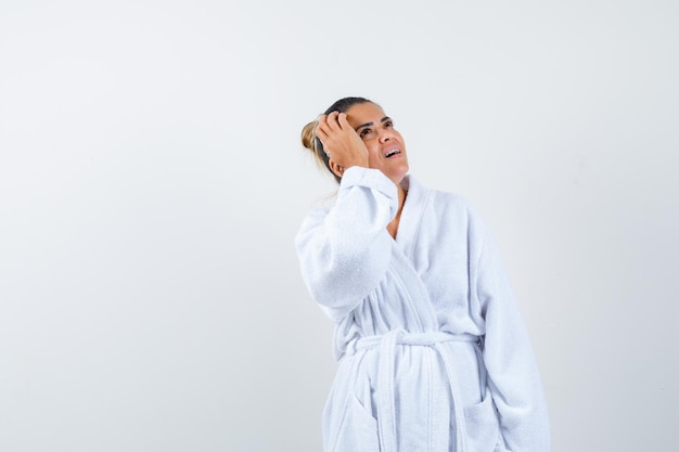 Free photo young woman holding hand on head while looking up in bathrobe and looking thoughtful