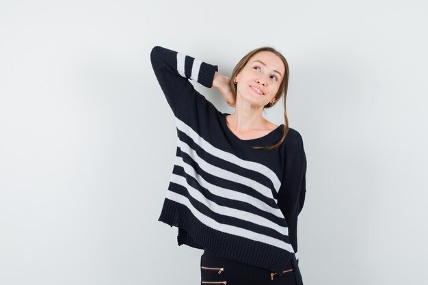 Young woman holding hand behind neck in striped knitwear and black pants and looking happy