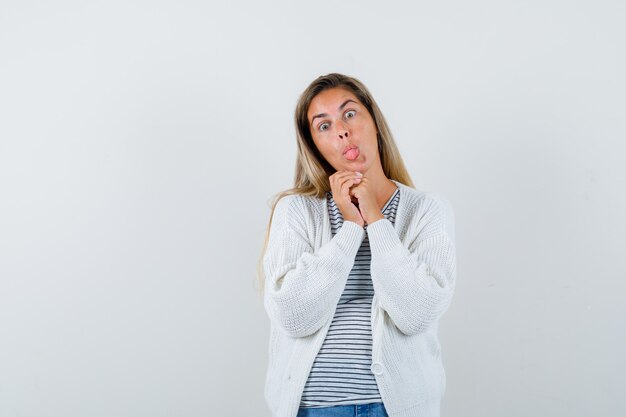 Young woman holding hands clasped, sticking out tongue in t-shirt, jacket and looking funny , front view.