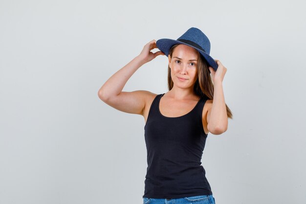 Young woman holding hands on hat and smiling in singlet, shorts front view.