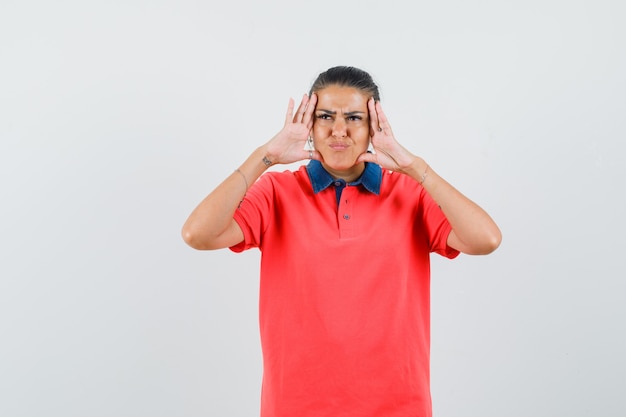 Young woman holding hands on head, grimacing in red t-shirt and looking dismal. front view.