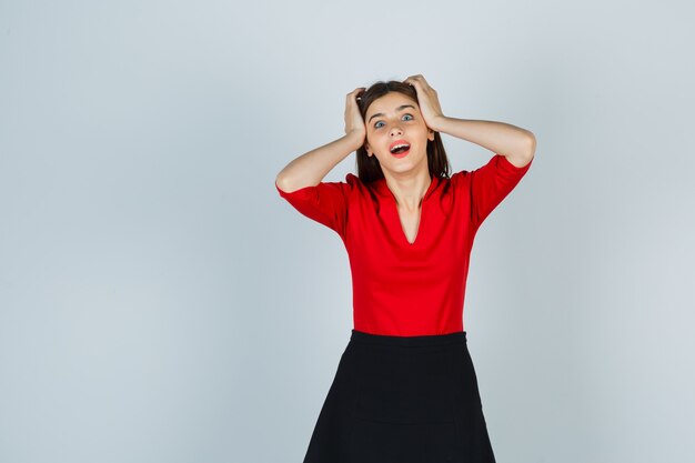 Young woman holding hands on head in red blouse, black skirt and looking surprised