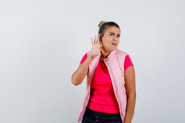 Young woman holding hands near ear to hear something in pink t-shirt and jacket and looking focused