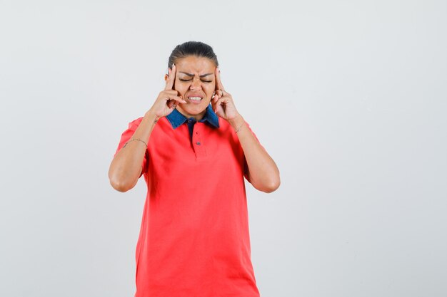 Young woman holding hands near head, having headache in red t-shirt and looking exhausted. front view.