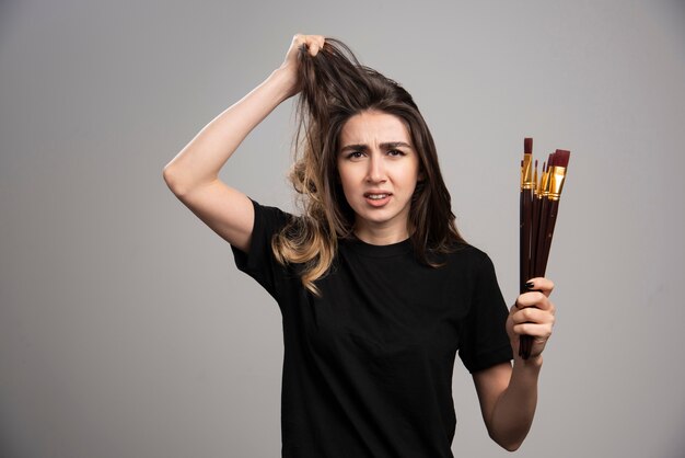 Young woman holding paint brushes touching her hair on gray wall.