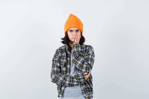 Young woman holding palm on cheek in orange hat checkered shirt looking pensive