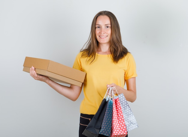 Young woman holding paper bags and cardboard box in yellow t-shirt, pants and looking glad