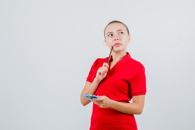 Young woman holding pencil and clipboard in red t-shirt and looking pensive