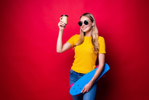 Young woman holding skateboard on shoulder and coffee isolated against red wall