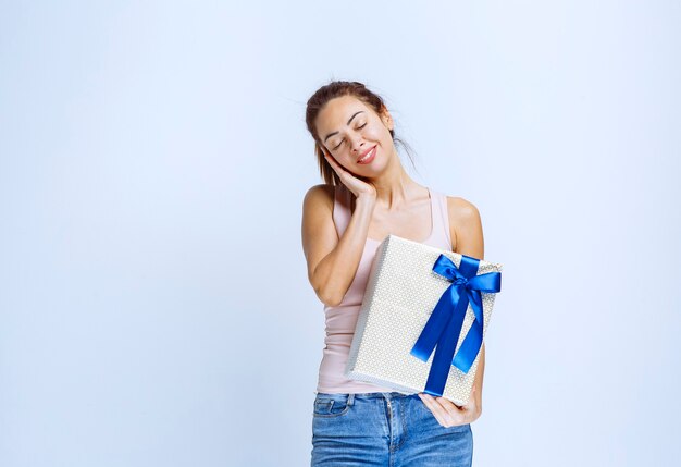 Young woman holding a white gift box wrapped with blue ribbon and looks tired and exhausted