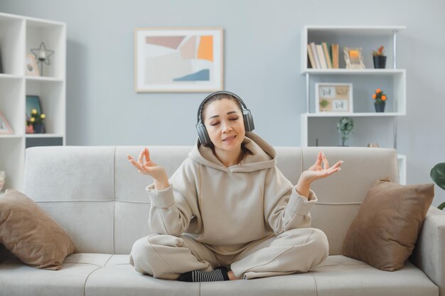 Young woman in home clothes sitting on a couch at home interior with headphones happy and positive relaxing meditating making meditation gesture with fingers