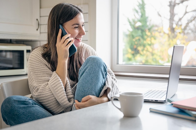 A young woman is sitting in the kitchen with a smartphone and a laptop