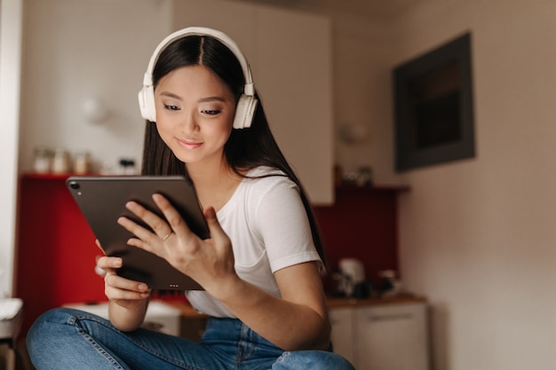 Free photo young woman in jeans and white t-shirt looks into tablet and listens to music in headphones