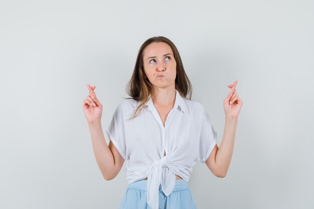 Young woman keeping fingers crossed in white blouse and light blue skirt and looking pensive