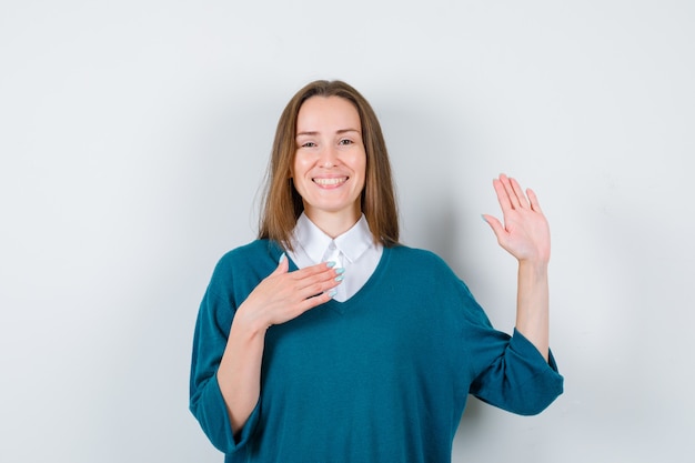 Young woman keeping hand on chest, showing palm in sweater over white shirt and looking merry. front view.