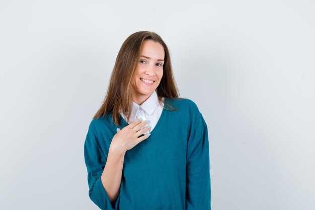 Young woman keeping hand on chest in sweater over white shirt and looking cheerful. front view.