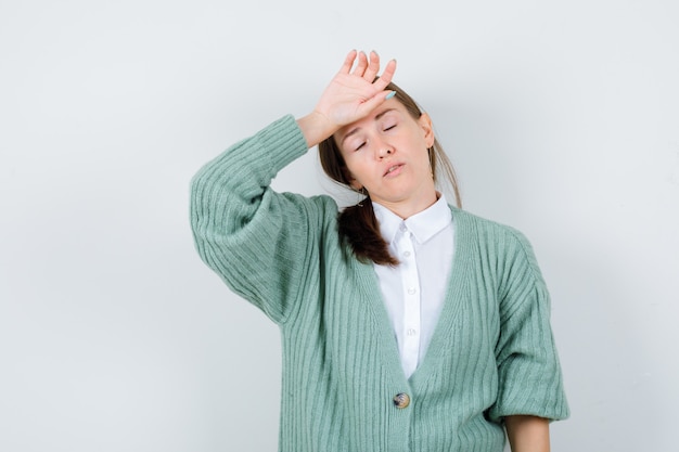 Young woman keeping hand on forehead, shutting eyes in blouse, cardigan and looking fatigued , front view.