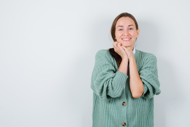 Young woman keeping hands under chin in blouse, cardigan and looking merry , front view.