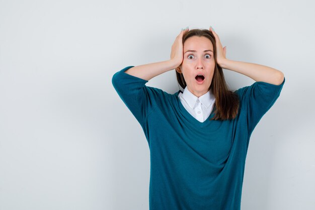 Young woman keeping hands on head in sweater over white shirt and looking shocked , front view.