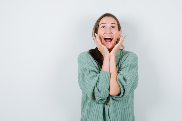 Young woman keeping hands near face while screaming, looking up in blouse, cardigan and looking blissful. front view.