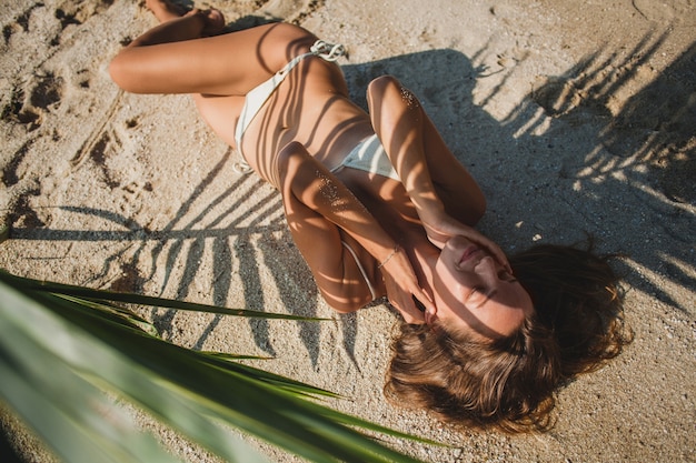 Free photo young woman laying on sand beach under palm tree leaf