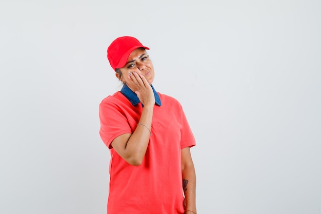 Young woman leaning cheek on palm, having toothache in red shirt and cap and looking exhausted. front view.