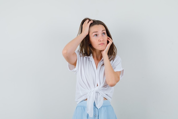 Young woman leaning cheek on palm while scratching head in white blouse and light blue skirt and looking cheerful