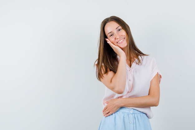 Young woman leaning cheek on raised palm in t-shirt, skirt and looking cheerful , front view.