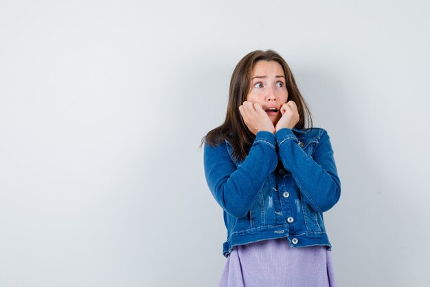 Young woman leaning cheeks on hands in t-shirt, jacket and looking terrified , front view.