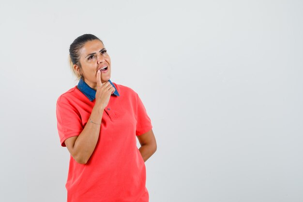 Young woman leaning finger near mouth, standing in thinking pose in red t-shirt and looking pensive. front view.