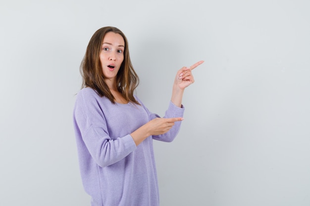 Young woman in lilac blouse pointing aside and looking interested 