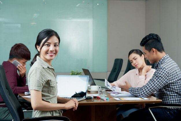 Young woman looking at camera distracted from the departmental meeting with colleagues