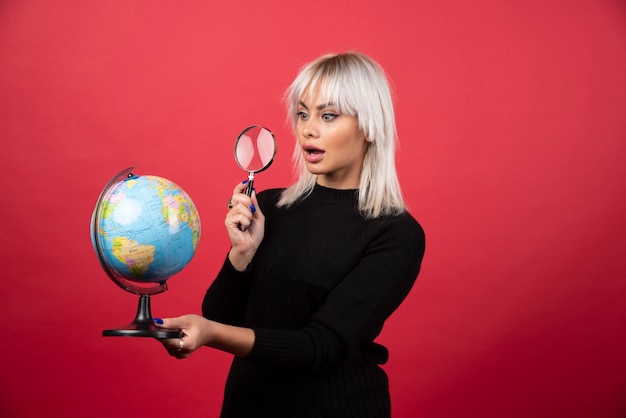 Young woman looking on a globe with loupe on a red background