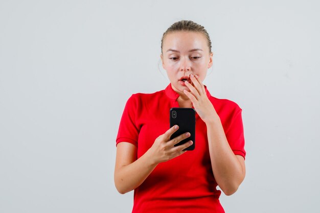 Young woman looking at mobile phone in red t-shirt and looking surprised