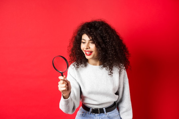 Young woman looking through magnifying glass investigating or searching for something stare curious ...