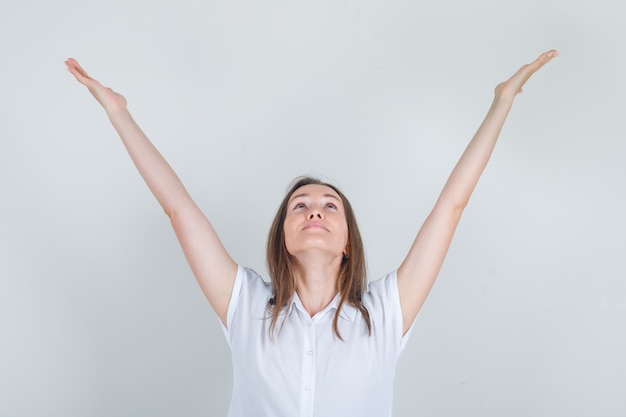 Free photo young woman looking up with raised hands in white t-shirt and looking cheerful