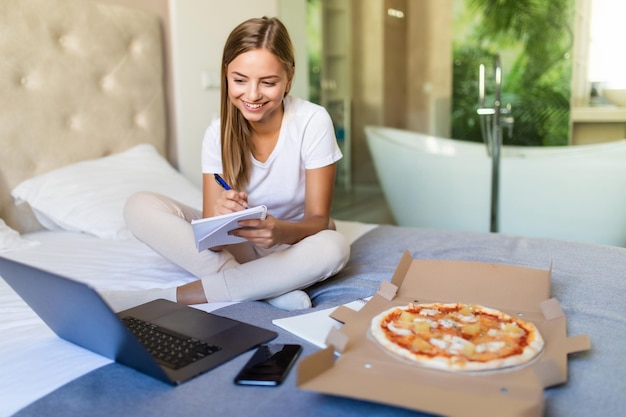 Free photo young woman lying on a bed in bedroom indoors at home while eating pizza and using laptop computer