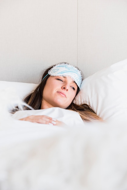 Young woman lying on bed with blanket wearing eye mask on her forehead