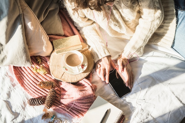 Free photo young woman lying on bed with cup of coffee and using smartphone