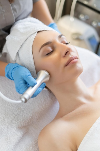 Free photo young woman lying on cosmetologist's table during rejuvenation procedure. cosmetologist take care about neck and face skin youthfull and wellness. hardware face cleaning procedure