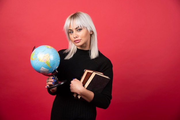 Free photo young woman model posing with a books and a earth globe on a red wall.