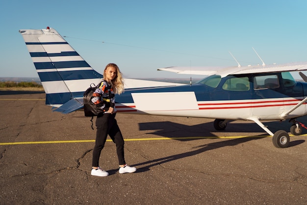 Young woman model with a modern haircut posing near a private plane wearing trendy casual outfit and black backpack